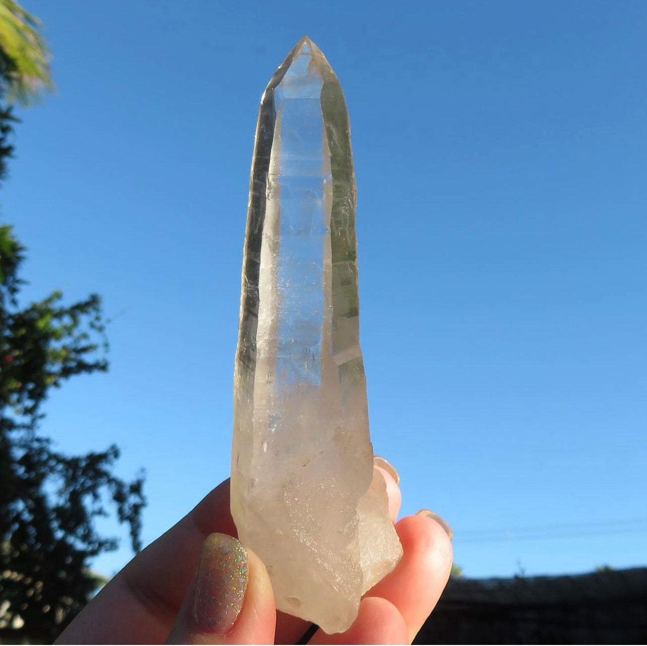 large optical lemurian quartz point with striations and etchings shining in the sunlight against bright blue sky. 